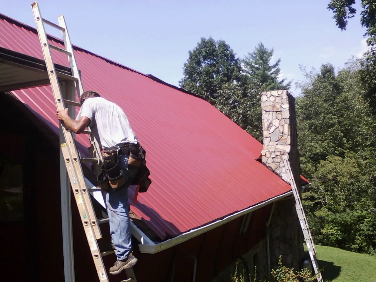 Roofer on a ladder beside a red metal roof and stone chimney — Soffit & Fascia Repair by Roofing Brothers in Nooksack